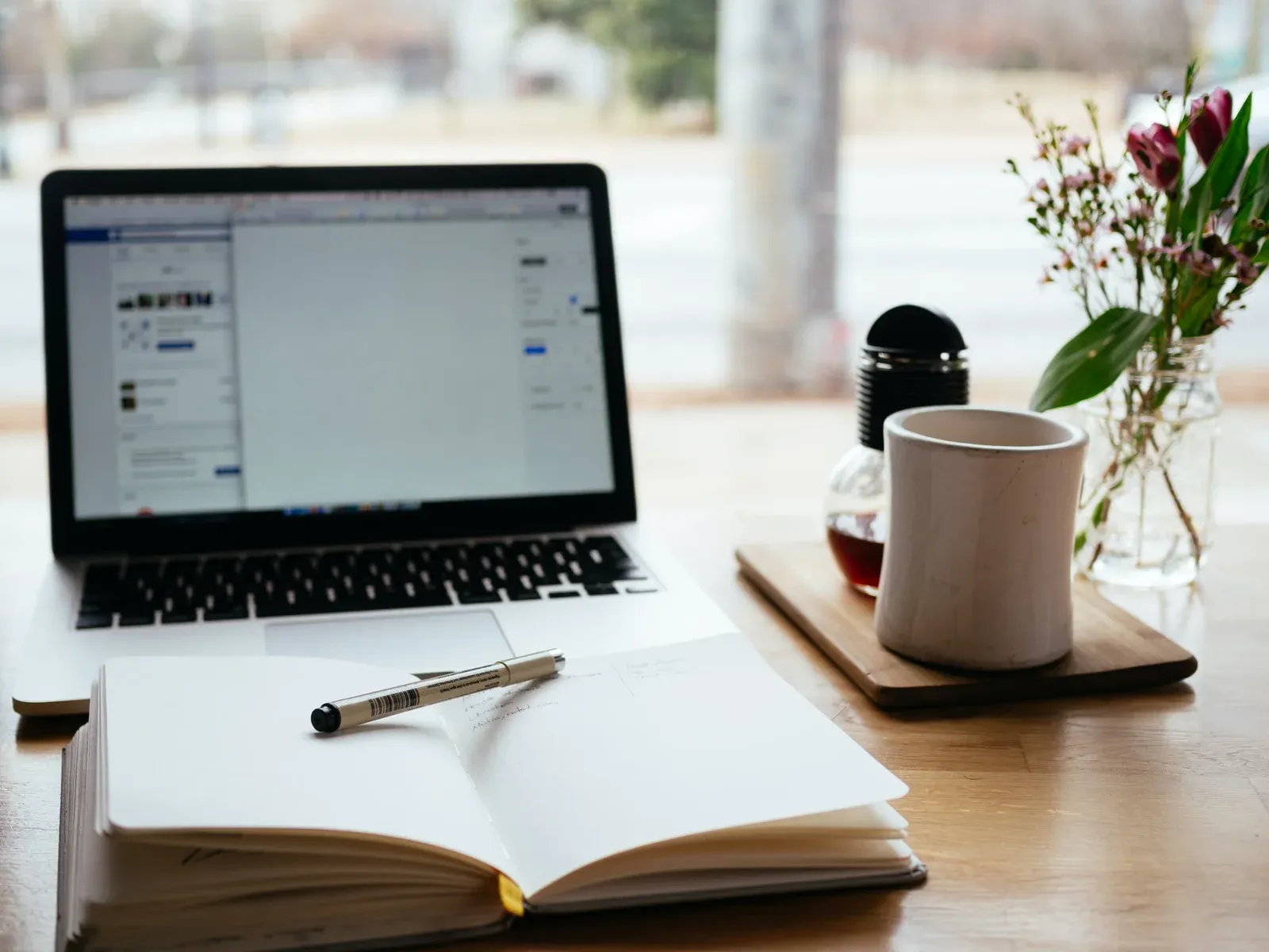 A laptop on a desk beside an open notebook and a cup